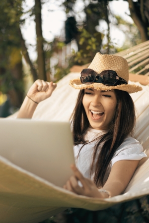 Eine Frau mit Strohhut und Sonnenbrille auf dem Kopf sitzt lässig in einer Hängematte, hat einen Laptop auf den Beinen liegen und freut sich über das Ergebnis - vielleicht ja ihren Newsletter Start, hehe - das sie darauf sieht