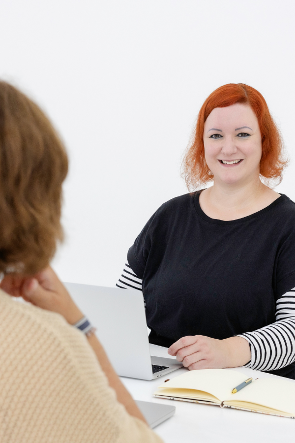 Stefanie sitzt mit dem Laptop und einem Notizbuch-Planer vor sich lächelnd einer Kundin gegenüber