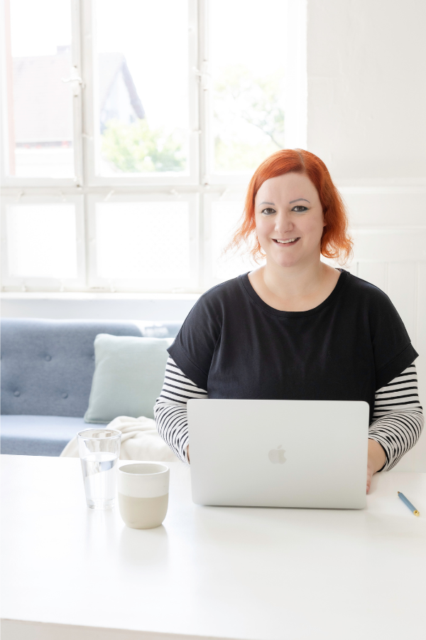 Stefanie sitzt freundlich lächelnd an einem Tisch, vor ihr steht ihr aufgeklappter Laptop